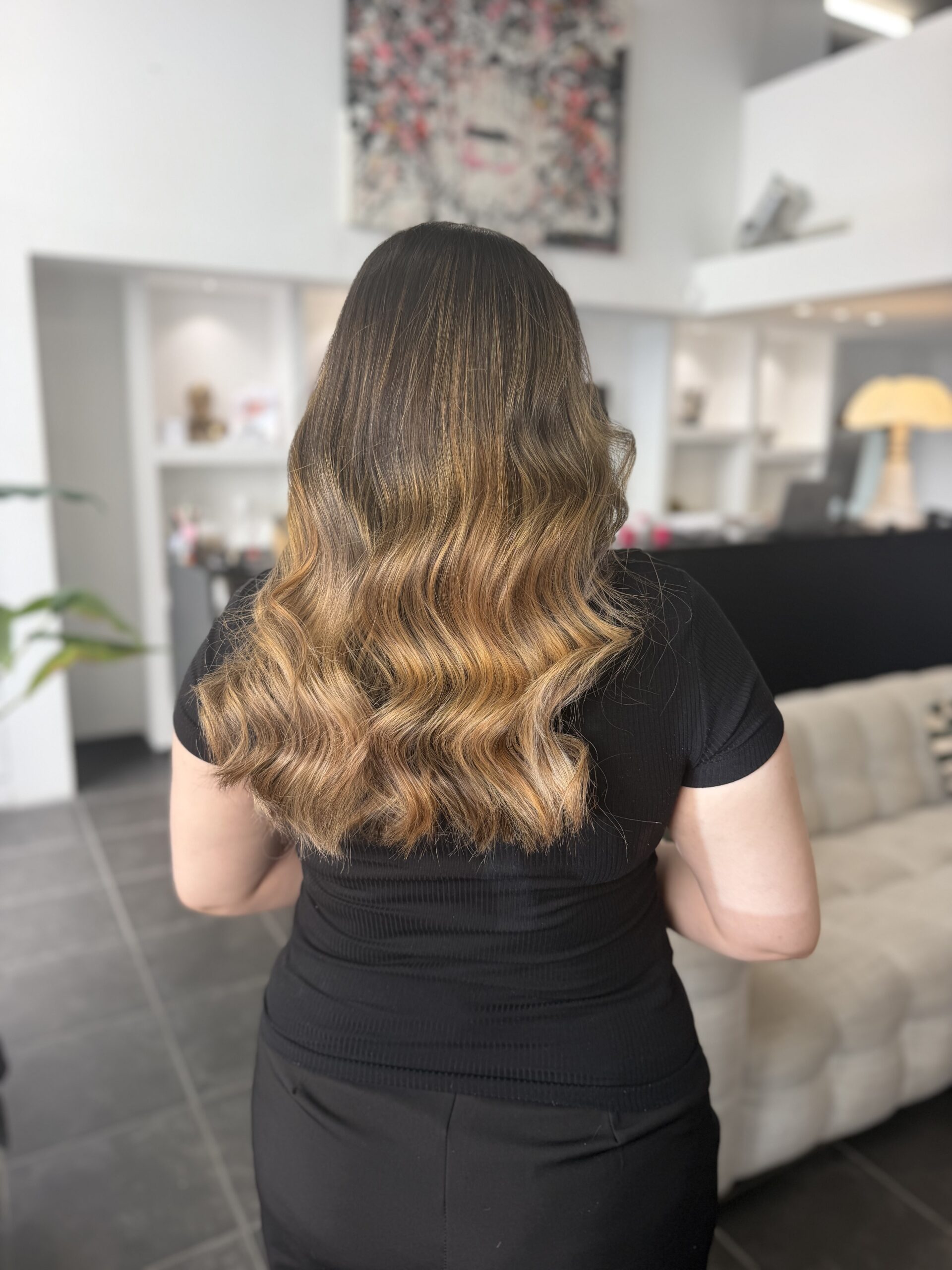 Vue de dos de cheveux mi-longs bruns, balayage dégradé miel et caramel, larges ondulations régulières