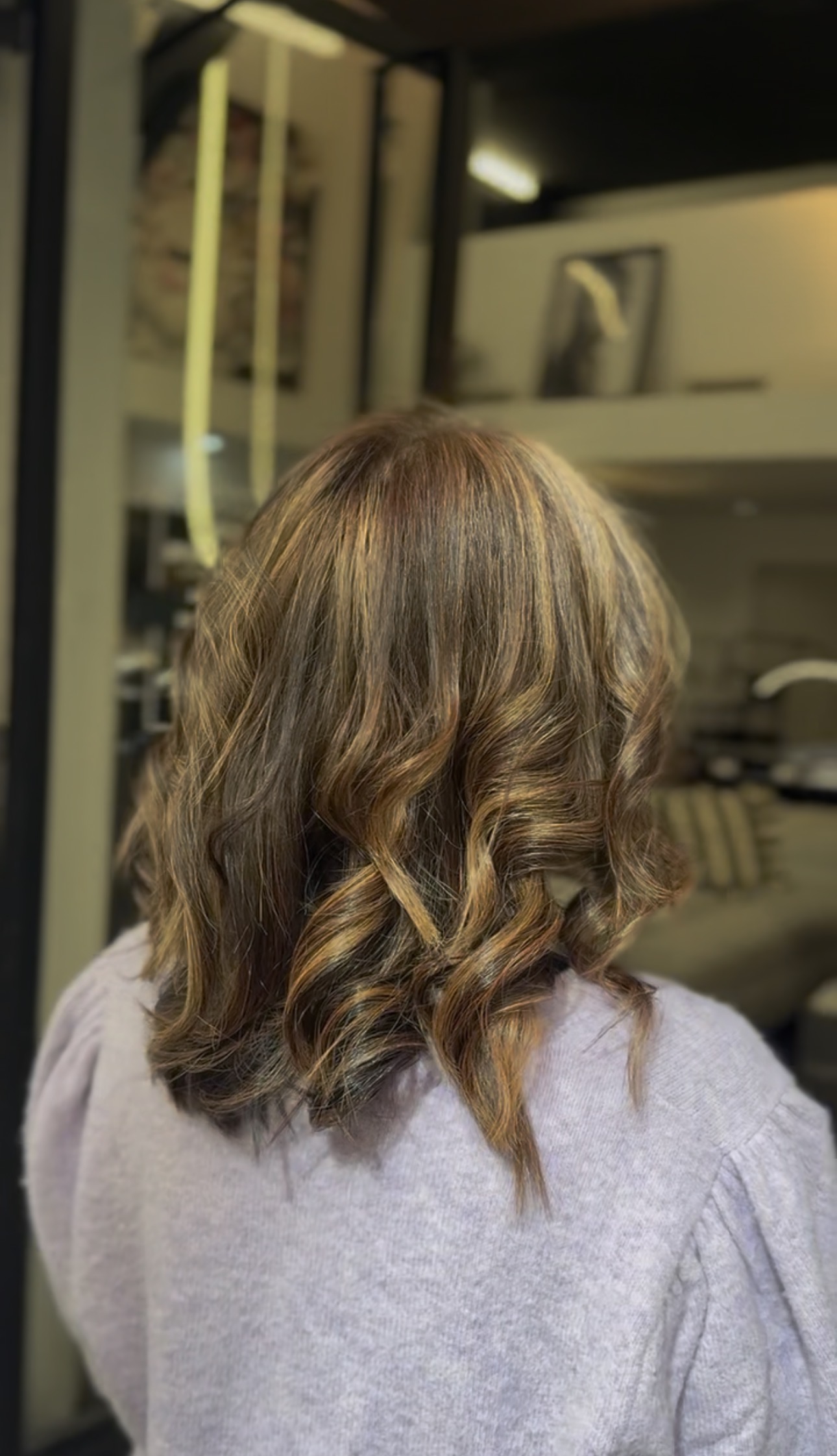 Vue de dos d'une coupe mi-longue aux pointes ondulées, balayage caramel et doré sur base châtain