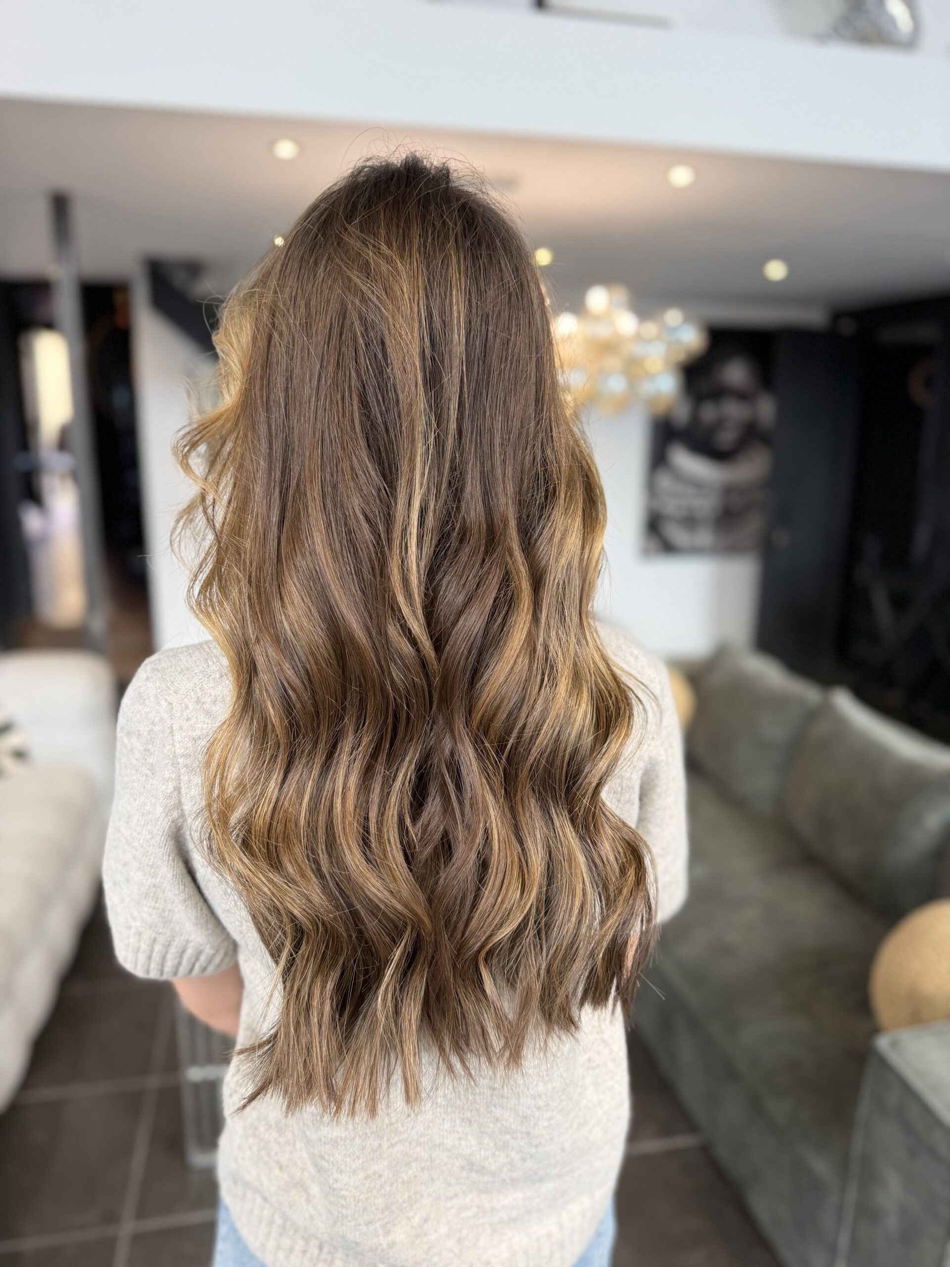 Vue de dos de cheveux longs ondulés, balayage caramel et miel sur base brune, dans un salon lumineux