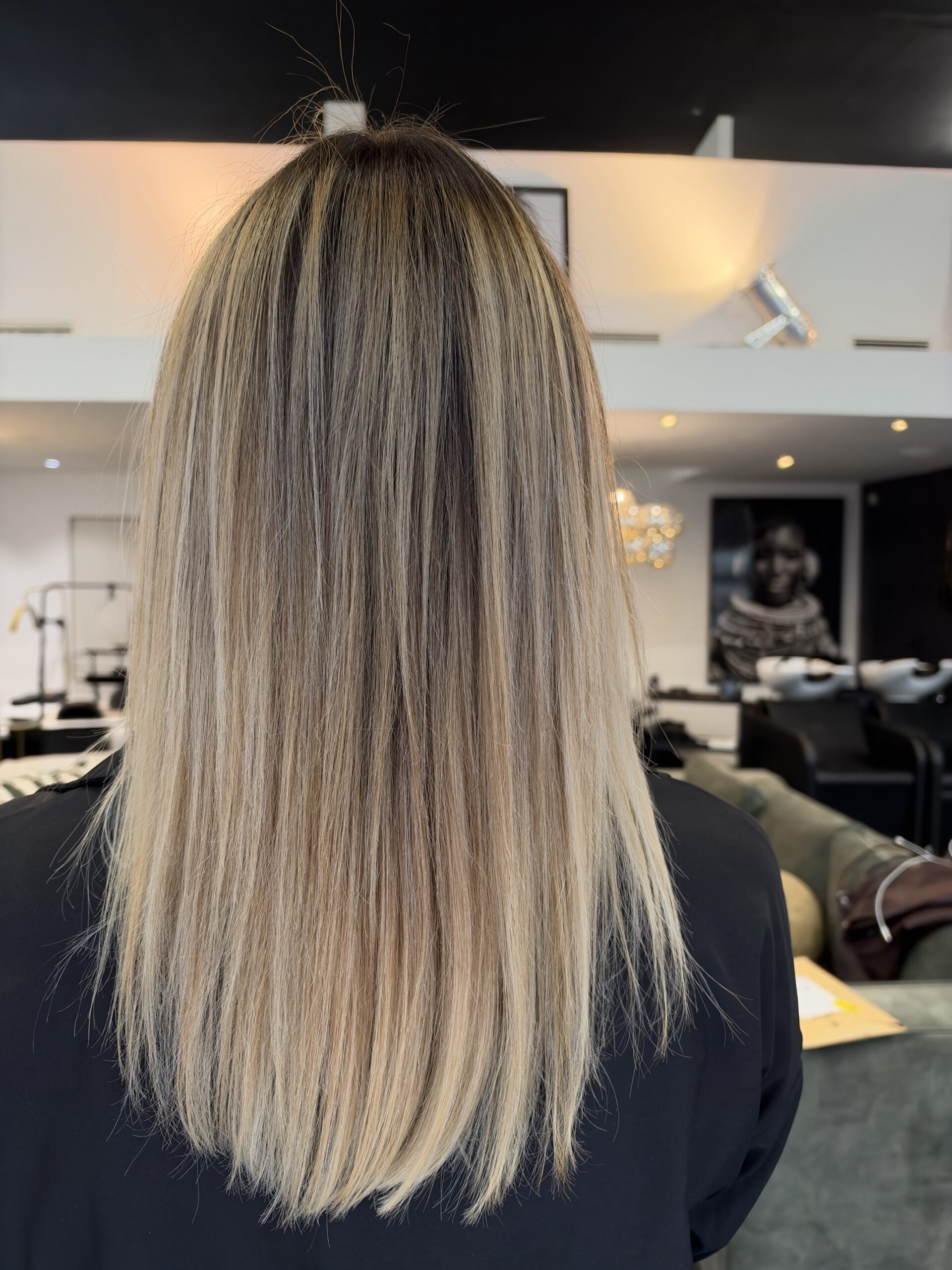 Vue de dos de cheveux longs et lisses, balayage blond cendré et beige sur racines châtain clair, dans un salon lumineux