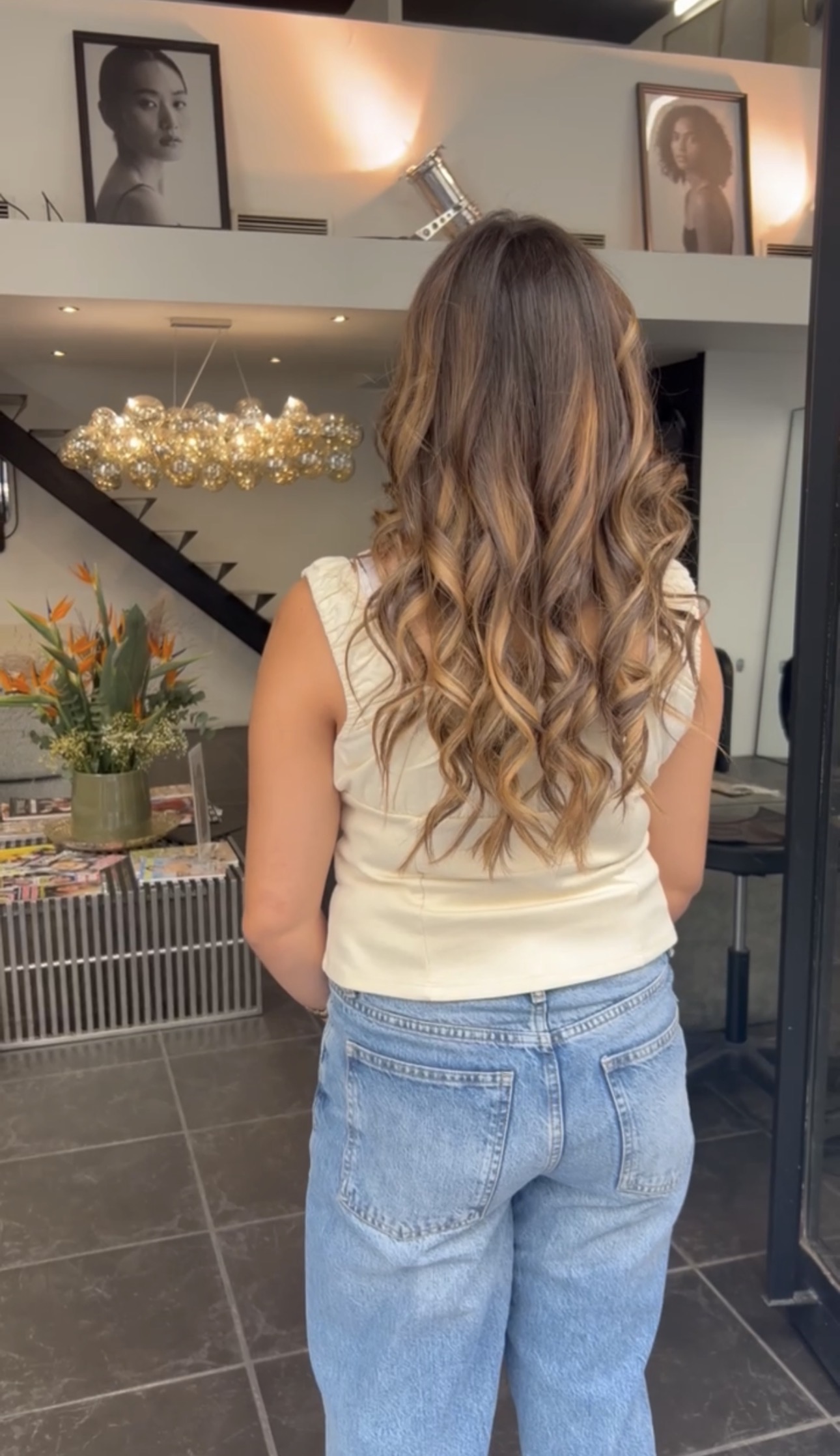 Vue de dos de cheveux longs bouclés, balayage caramel et blond doré sur base brune, dans le salon