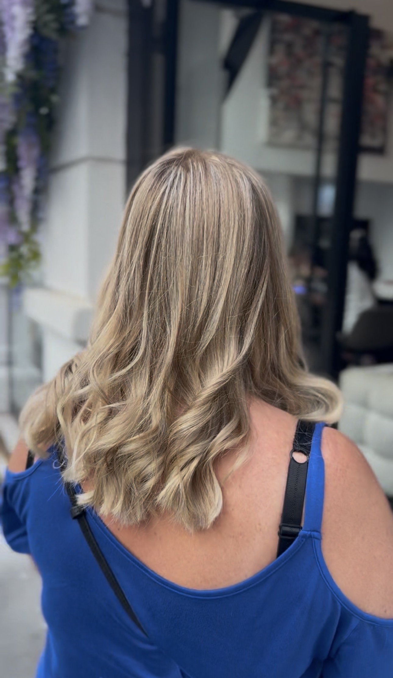 Vue de dos de cheveux mi-longs, balayage blond lumineux et ondulations souples, devant l'entrée du salon