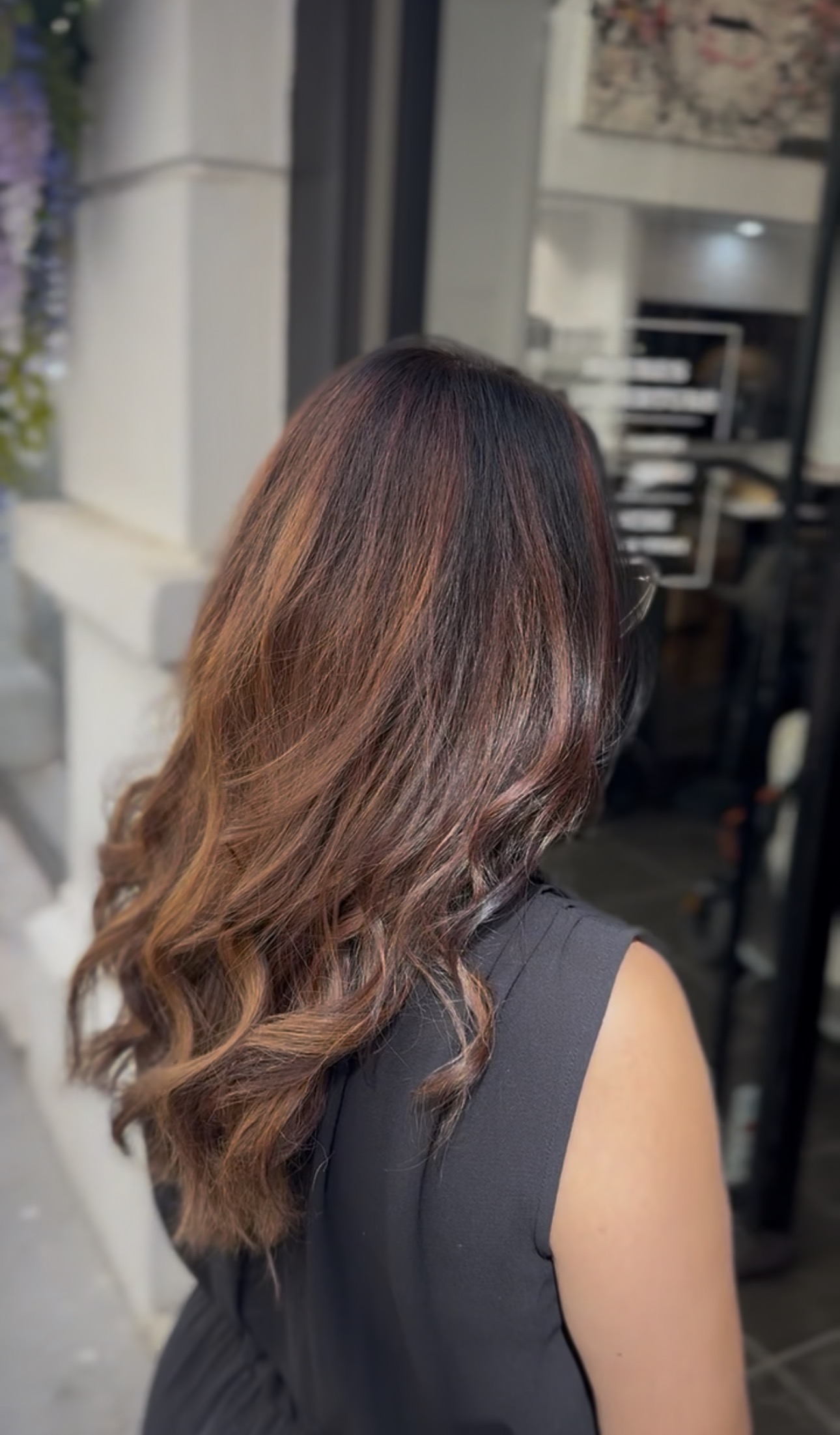 Vue de dos de cheveux longs ondulés, balayage brun chaud et caramel devant l'entrée du salon