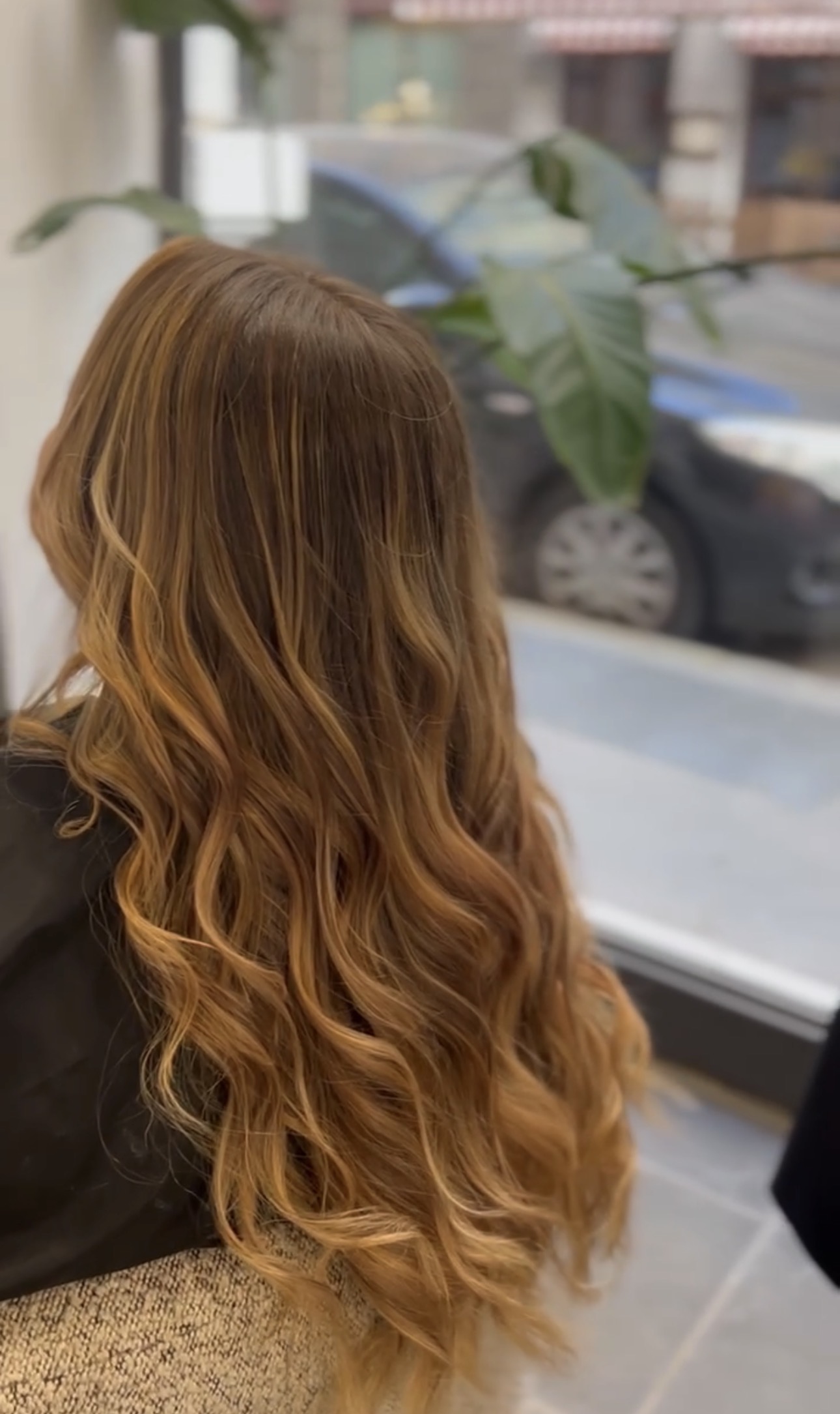 Vue de dos de cheveux très longs, balayage caramel et miel sur base châtain cendré, larges boucles, devant la vitrine du salon
